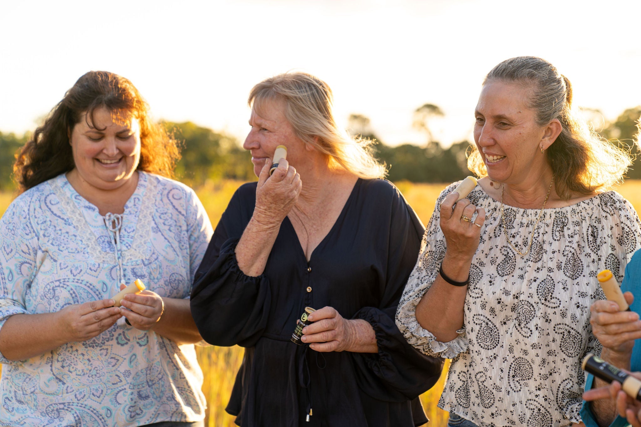 three mature women smiling and walking in a field, applying face balm to their skin