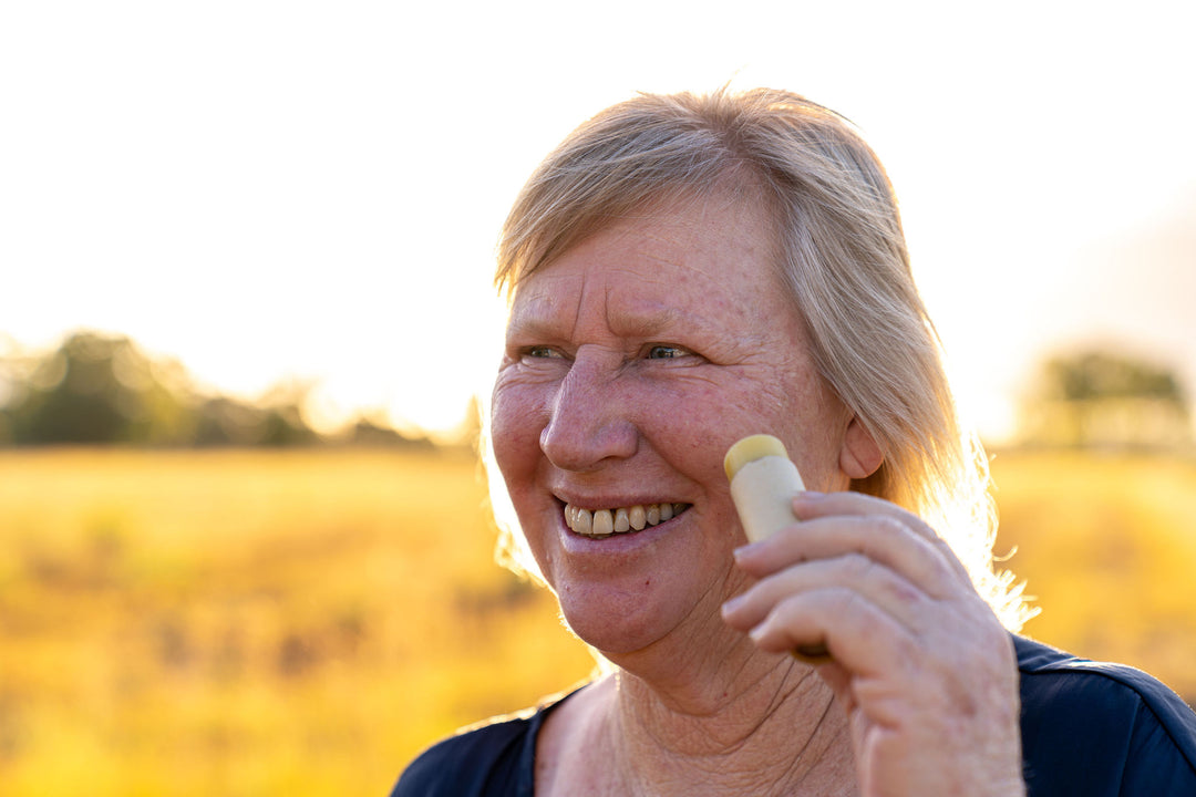 The Founder of Australian Hand and Skin balm brand Mel smiling and looking to the side of the camera. She is in a field and is holding a stick of face balm to her face